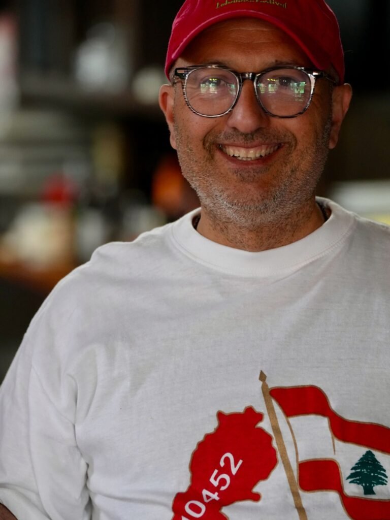 Elie, owner and founder of That Lebanese Place, smiling in the restaurant wearing a white T-shirt with the Lebanese flag and a red cap.