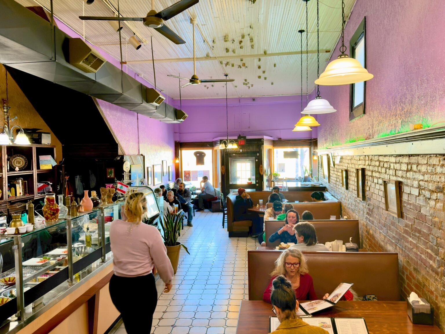 Lively dining room at That Lebanese Place with guests seated in booths enjoying Mediterranean dishes, warm lighting, exposed brick wall, and open counter service.