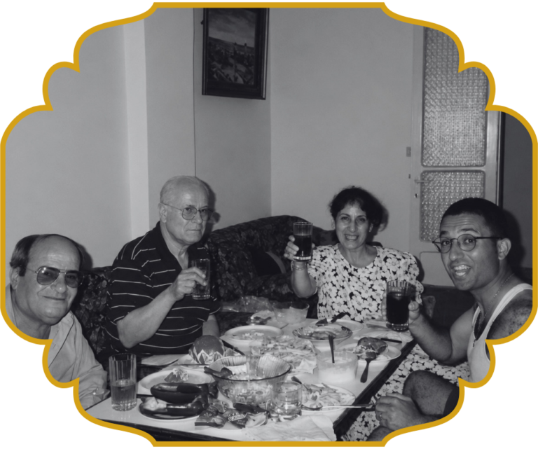 Four family members gathered around a home table in Lebanon, raising glasses in a toast over a spread of Lebanese dishes including stuffed vegetables, pita, and dips.