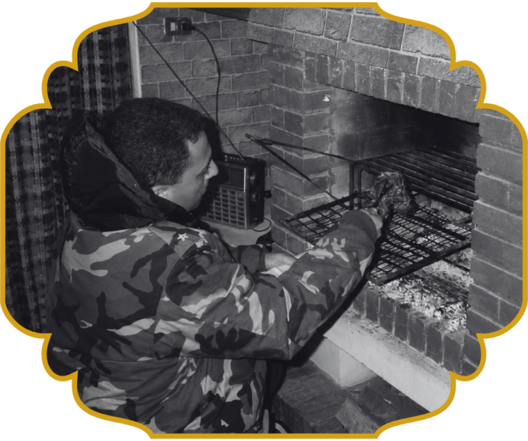 A young Chef Elie grilling meat over a wood fire in a traditional brick hearth in Lebanon, tending skewers on the grill grate.