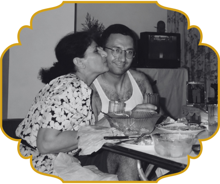 A woman kissing a smiling young Chef Elie on the cheek at a family meal in Lebanon, with bowls of food and flatbread on the table in front of them.