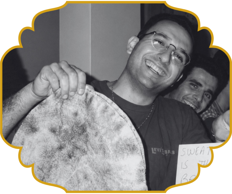 A young Chef Elie smiling and holding up a large round loaf of Lebanese flatbread, with a laughing friend beside him.