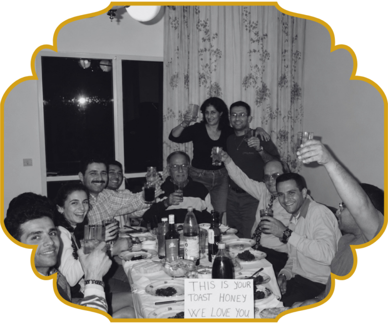 A large family gathered around a festive dinner table in Lebanon, everyone raising their glasses in a toast, with a handwritten sign on the table reading "This is your toast honey, we love you."